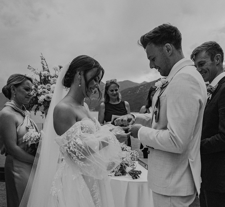 Ein stimmungsvolles Schwarz-Weiß-Foto, das den Moment des Ringtauschs während einer Hochzeitszeremonie im Freien einfängt. Der Bräutigam in einem hellen Anzug steckt der freudig lächelnden Braut den Ehering an den Finger. Die Braut trägt ein romantisches Hochzeitskleid mit auffälligen, schulterfreien und voluminösen Tüllärmeln, die mit feinen Blattmustern bestickt sind, sowie einen langen Schleier. Im Hintergrund stehen glückliche Hochzeitsgäste und Trauzeugen, die das Paar mit einem Lächeln beobachten. Dahinter ist eine sanfte Berglandschaft unter einem leicht bewölkten Himmel zu erkennen.